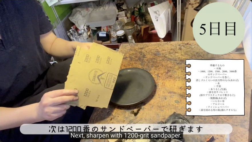 Progress through finer grits (1200 → 3000) — The maker presents the 1200-grit paper beside the evenly abraded lacquer surface before continuing through the finer grits.