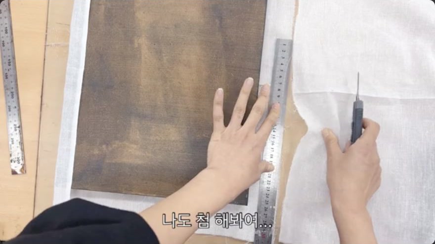 Cut the cloth to size — A ruler and knife guide a straight cut through the cloth, leaving a measured margin around the panel.